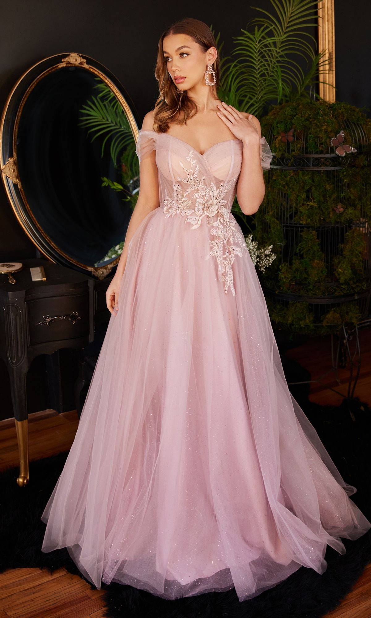 A woman stands indoors by a black vanity and large round mirror, elegantly wearing the Mauve Pink Long A-Line Prom Dress CD3394 with floral applique and an off-the-shoulder design.